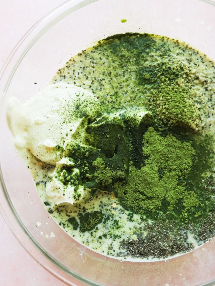 A glass bowl containing a mixture of green matcha powder, chia seeds, yogurt, and milk, before being stirred together.