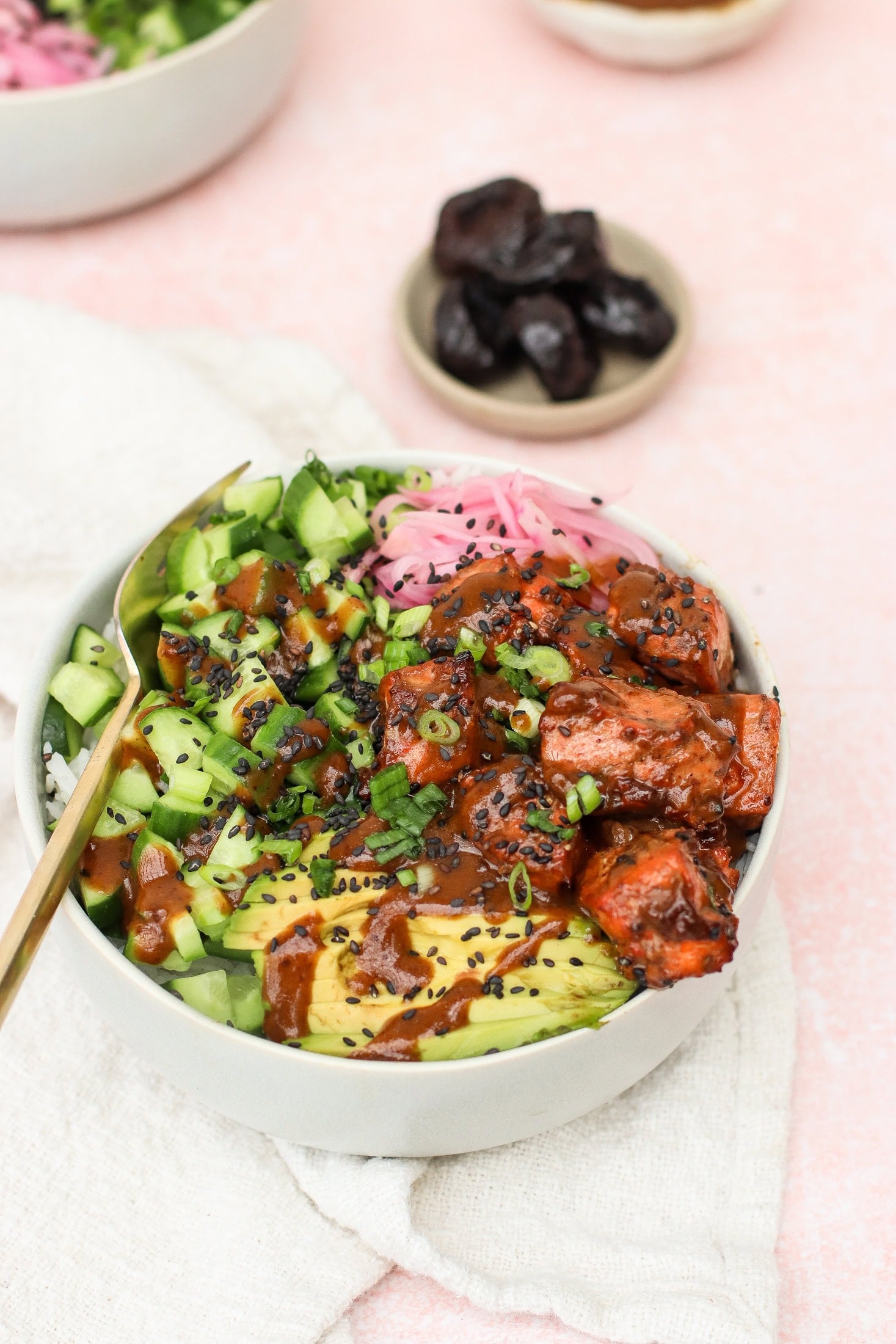 A bowl with sliced avocado, cucumber, marinated tofu, pickled onions, chopped scallions, black sesame seeds, and sauce, served on a light cloth with a small bowl of prunes in the background.