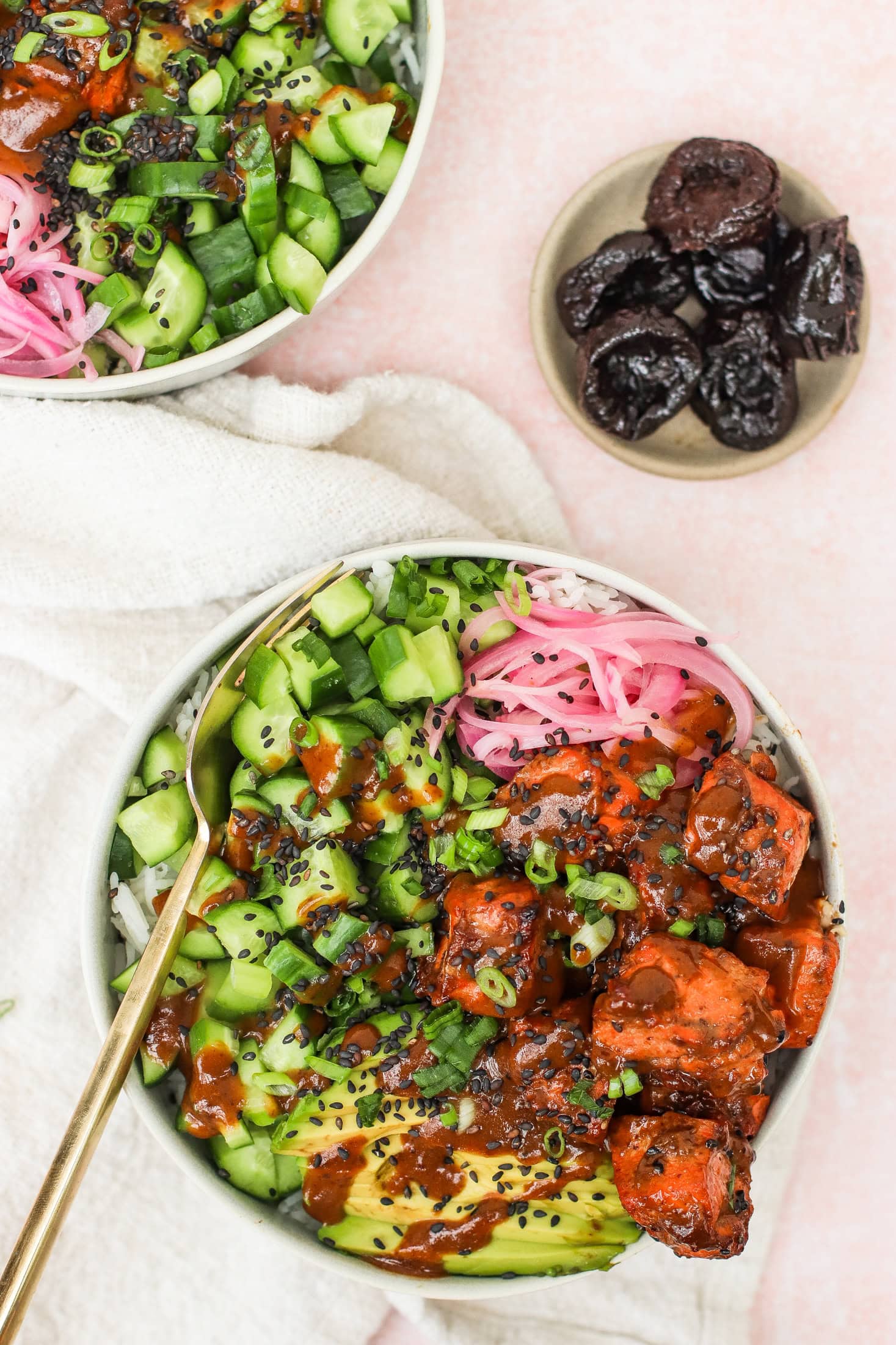 A bowl filled with rice, diced cucumber, pickled onions, avocado slices, marinated tofu, green onions, and black sesame seeds. A small plate of dried prunes sits nearby on a light pink surface.