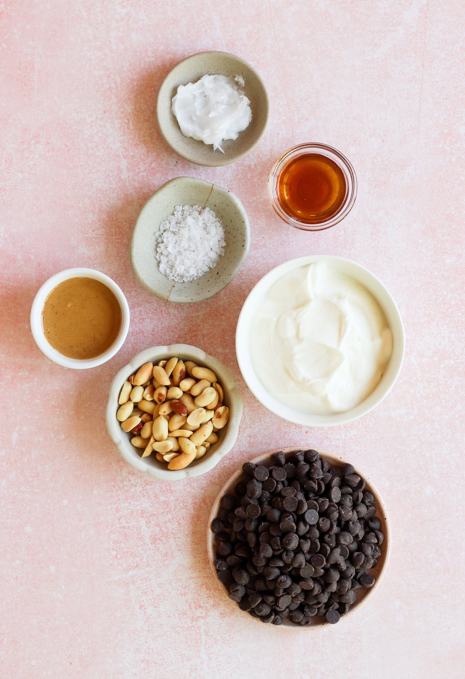 Seven small bowls on a pink surface, containing chocolate chips, peanuts, creamy yogurt, almond butter, coconut oil, flaky sea salt, and a small glass of vanilla extract.