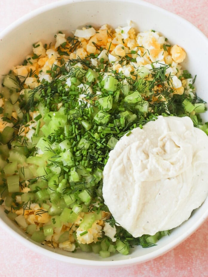 A white bowl filled with chopped hard-boiled eggs, celery, fresh dill, chives, and a large dollop of creamy mayonnaise or dressing, all ready to be mixed together.