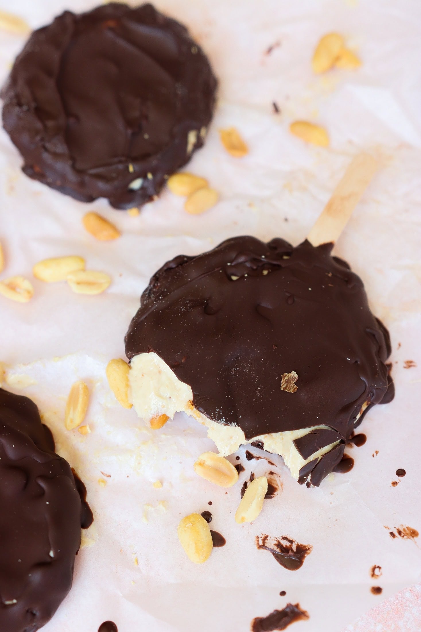 Chocolate-covered ice cream pops with peanuts scattered around. One pop has a bite taken out, revealing creamy ice cream inside. The treats are on white parchment paper with melted chocolate spots.