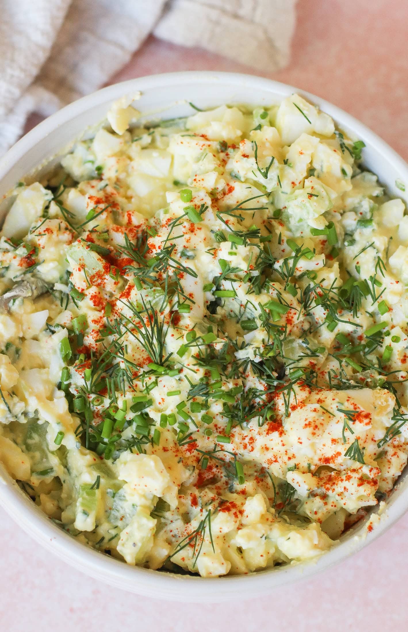 A bowl of creamy egg salad garnished with chopped chives, fresh dill, and a sprinkle of paprika, set on a light pink surface with a textured cloth in the background.
