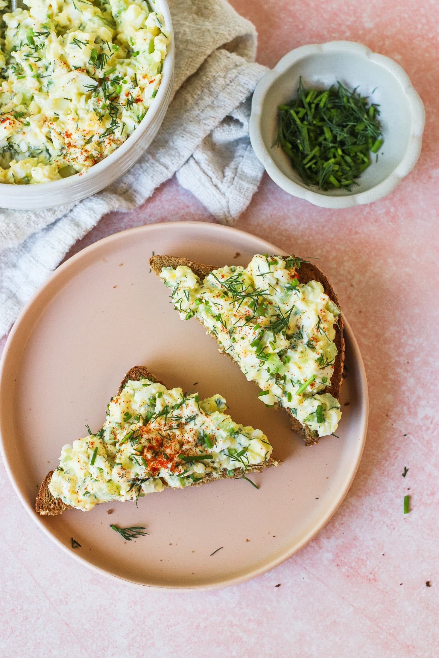 Two slices of bread topped with creamy egg salad and herbs are on a pink plate. Nearby are a bowl of egg salad, a small dish of chopped chives, and a light-colored cloth napkin on a pink surface.