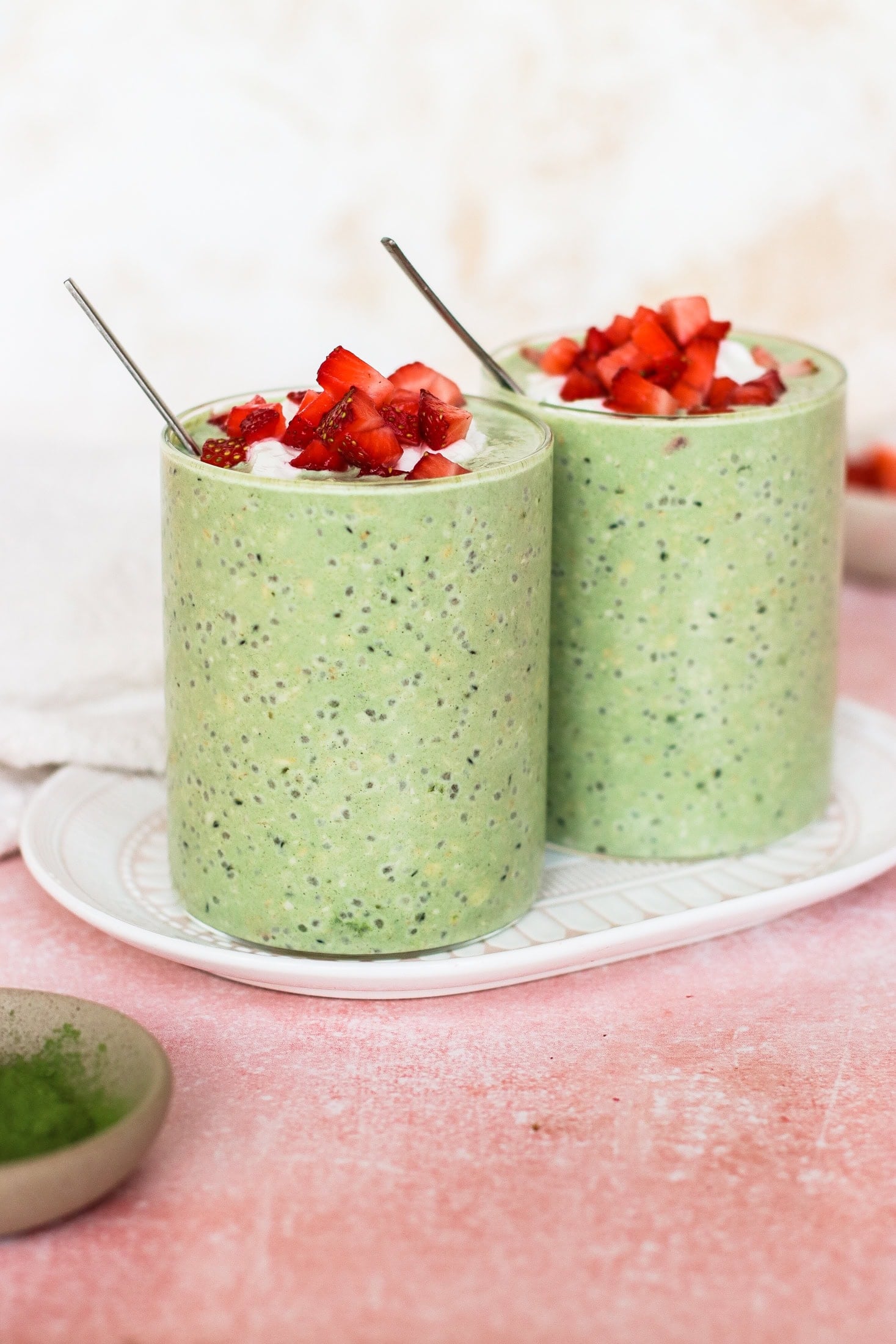Two glasses of green chia pudding topped with chopped strawberries and a spoon in each, placed on a white tray against a light pink background.