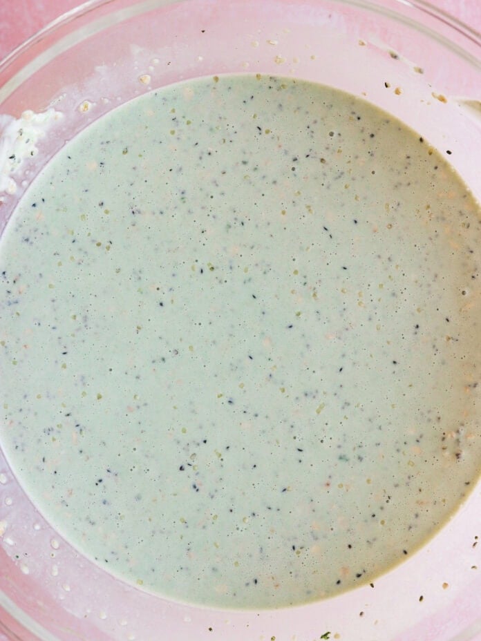 A clear glass bowl filled with a creamy, speckled dressing or sauce, viewed from above, set against a pink background.