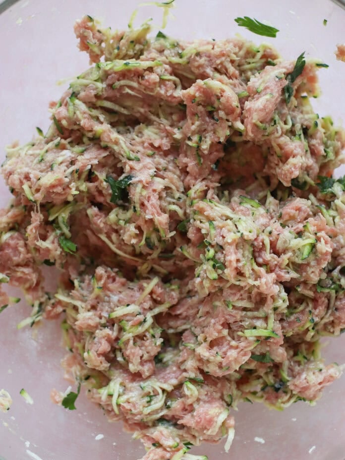 A close-up of raw ground meat mixed with shredded zucchini and herbs in a glass bowl, ready for cooking.