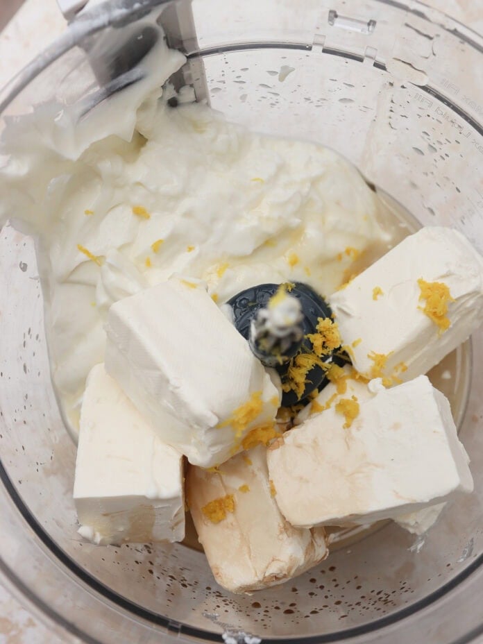A food processor bowl filled with cream cheese blocks, Greek yogurt, lemon zest, and a light brown liquid, ready to be blended.
