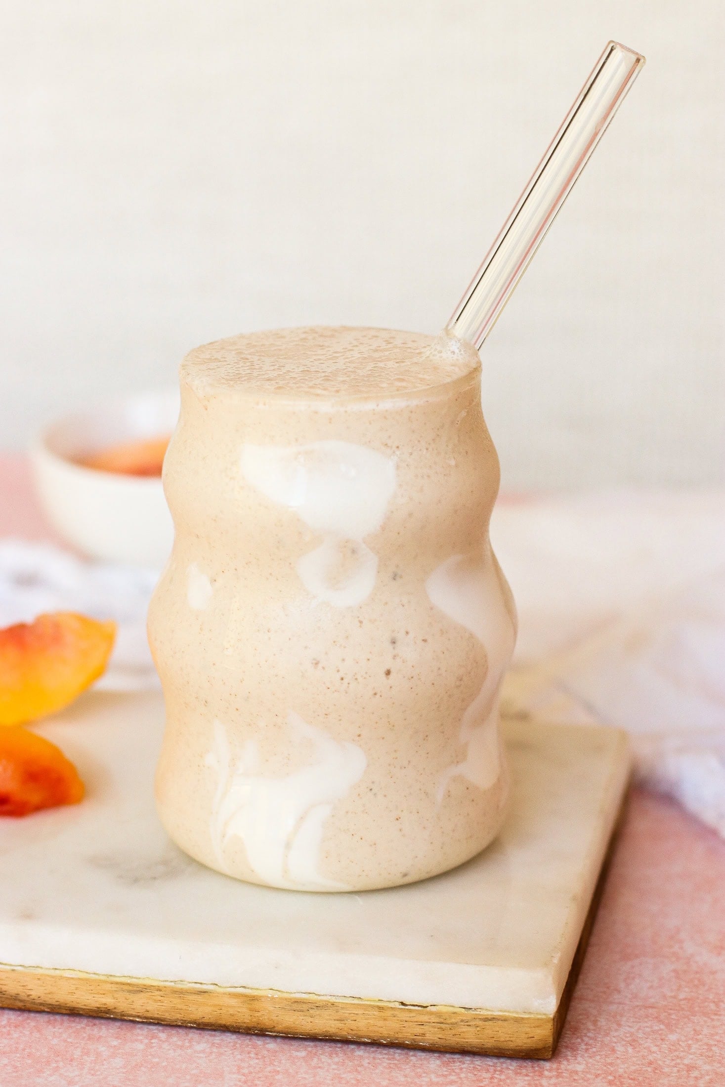 A creamy, light brown smoothie with white swirls is served in a wavy glass with a clear straw, placed on a marble board with peach slices nearby.