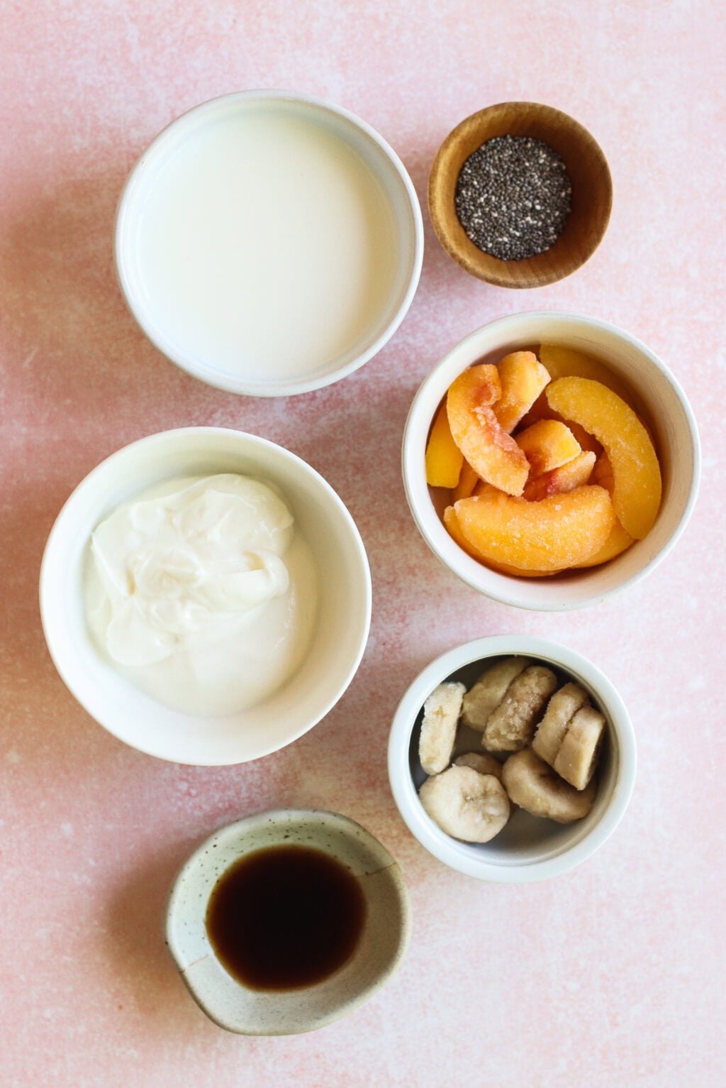 Six bowls and cups containing various ingredients are arranged on a pink surface. The ingredients include milk, chia seeds, frozen peach slices, yogurt, frozen banana slices, and vanilla extract.