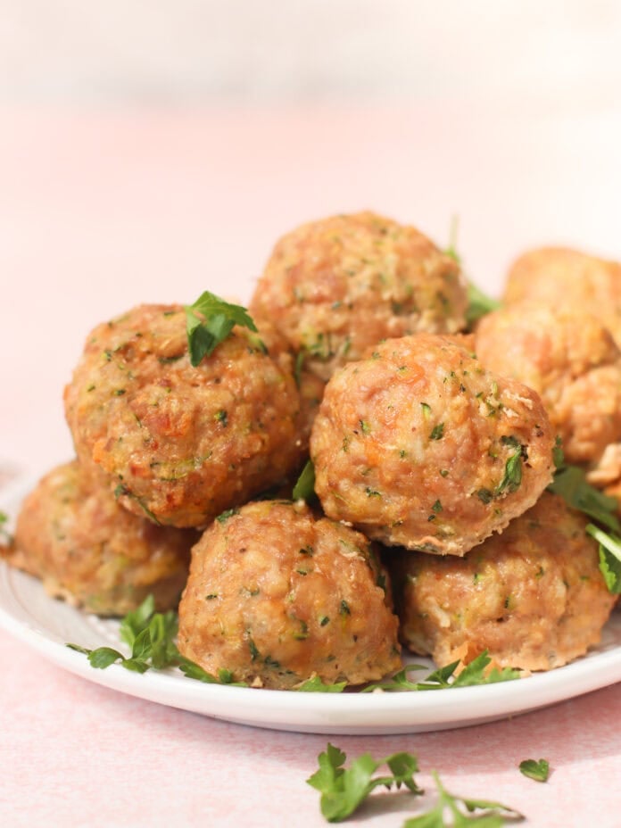 A plate stacked with golden-brown meatballs garnished with chopped parsley, set on a light pink surface with more parsley scattered around.