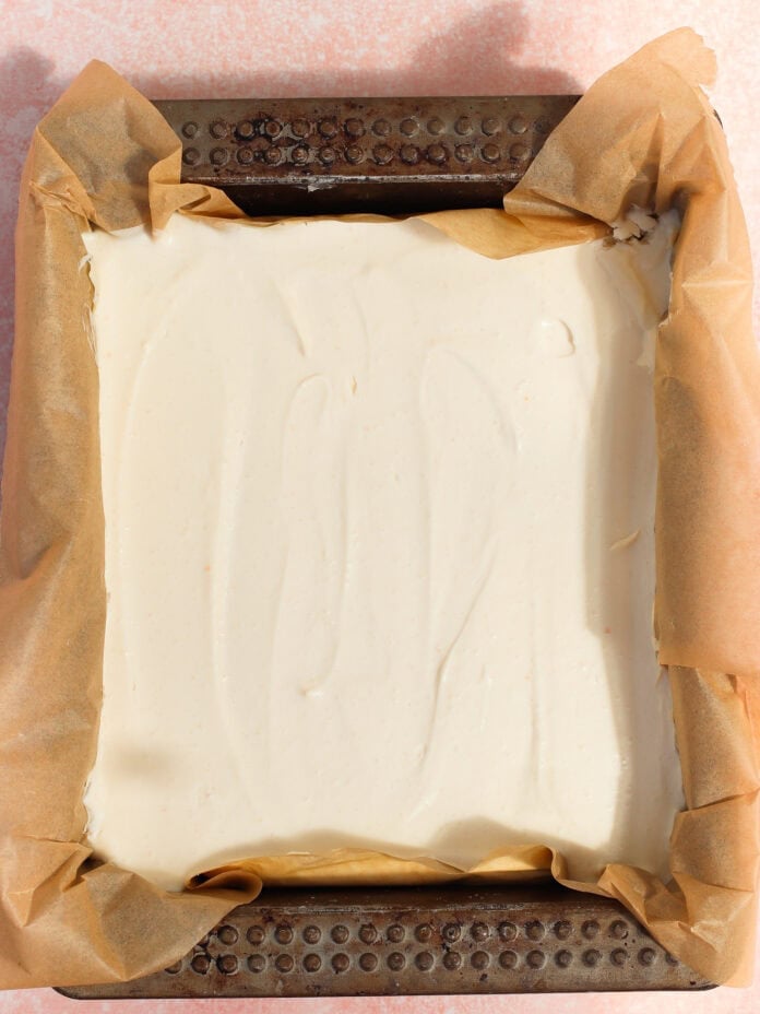 A square baking pan lined with brown parchment paper, filled with a smooth, creamy white batter or mixture, ready to be baked. The pan rests on a light pink surface.