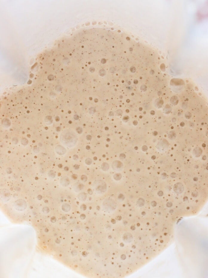 A close-up view of a creamy, bubbly batter or smoothie mixture inside a blender, with frothy bubbles scattered across the light beige surface.