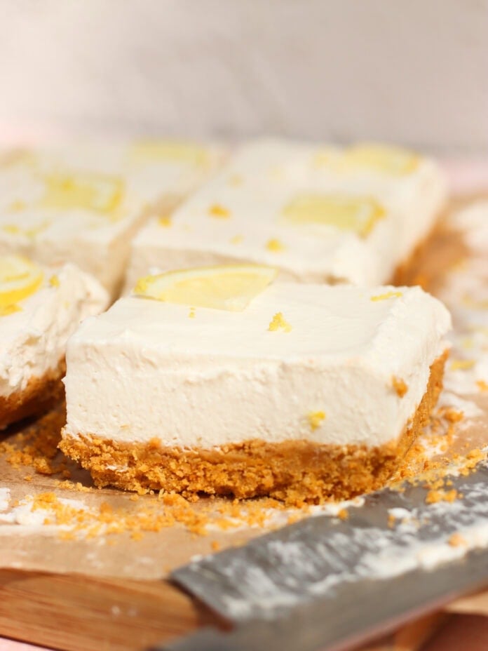 Close-up of creamy, no bake lemon cheesecake bars with a crumbly biscuit base, topped with thin lemon slices, on a wooden board. A knife with crumbs on its blade is in the foreground.