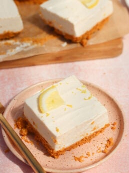 A square slice of creamy lemon cheesecake with a crumbly crust, topped with a lemon wedge, sits on a pink plate with a fork. More cheesecake pieces are visible in the background on parchment paper.