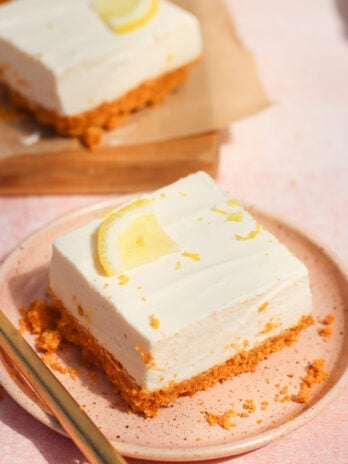 A square slice of creamy lemon cheesecake with a graham cracker crust, topped with a lemon wedge, served on a pink plate with a fork beside it. Another slice is in the background.