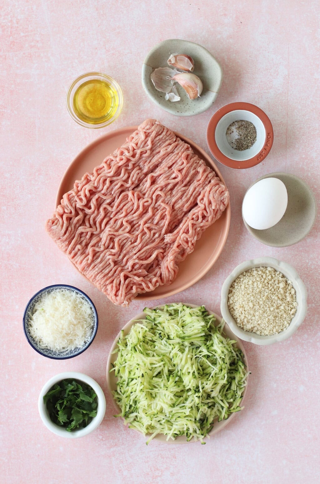 A flat lay of ground meat, shredded zucchini, grated cheese, an egg, breadcrumbs, chopped herbs, olive oil, garlic cloves, and seasonings in small bowls on a light pink surface.
