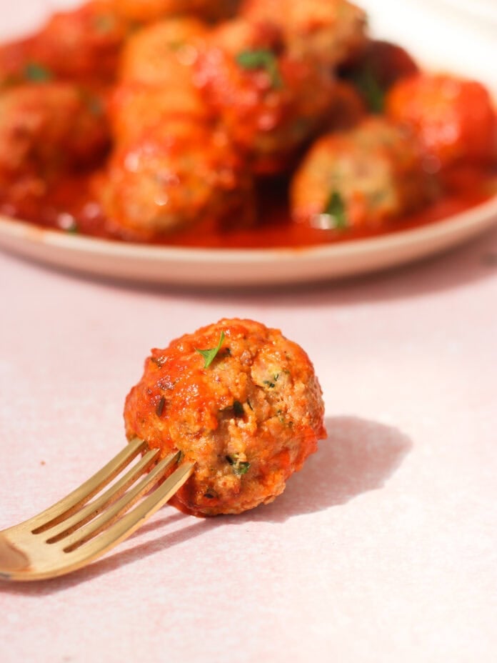 A golden fork holds a single meatball covered in tomato sauce, with a plate of more saucy meatballs blurred in the background on a light pink surface.
