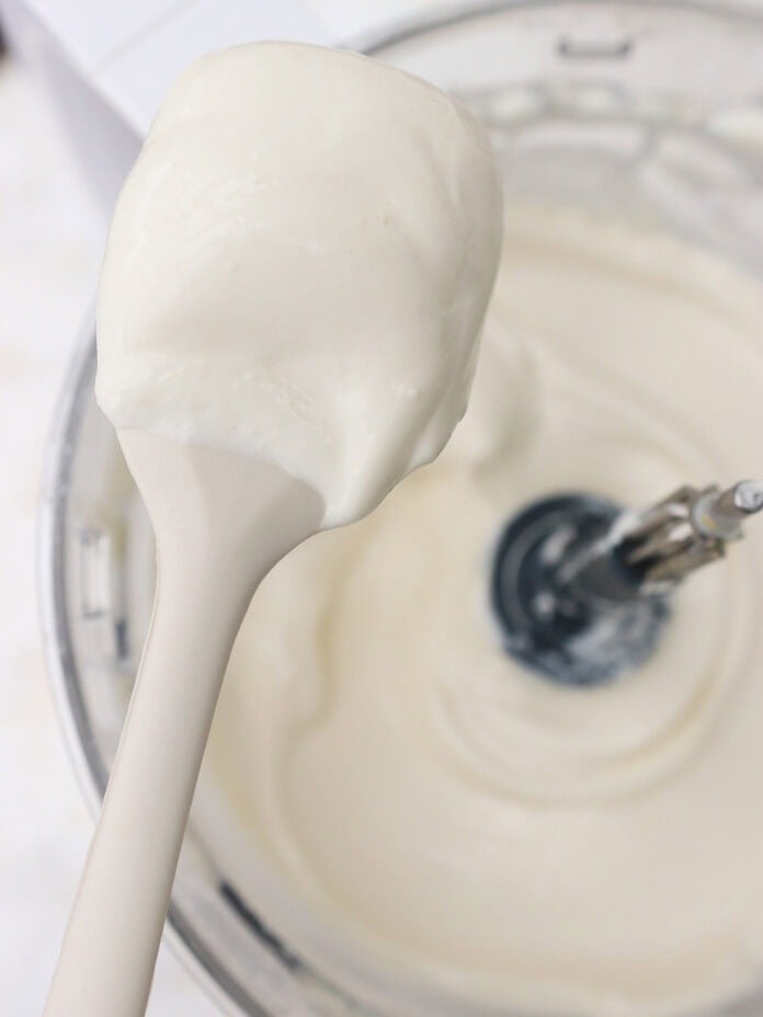 A close-up of a spoon holding thick, creamy white mixture above a mixing bowl with an electric mixer inside.
