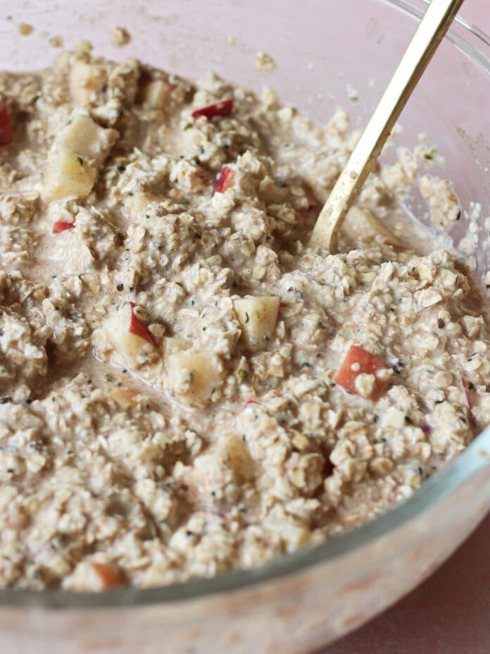 A glass bowl filled with a mixture of oats, chia seeds, and diced apples, being stirred with a gold spoon. The oat mixture appears creamy and thick, suggesting preparation for overnight oats.