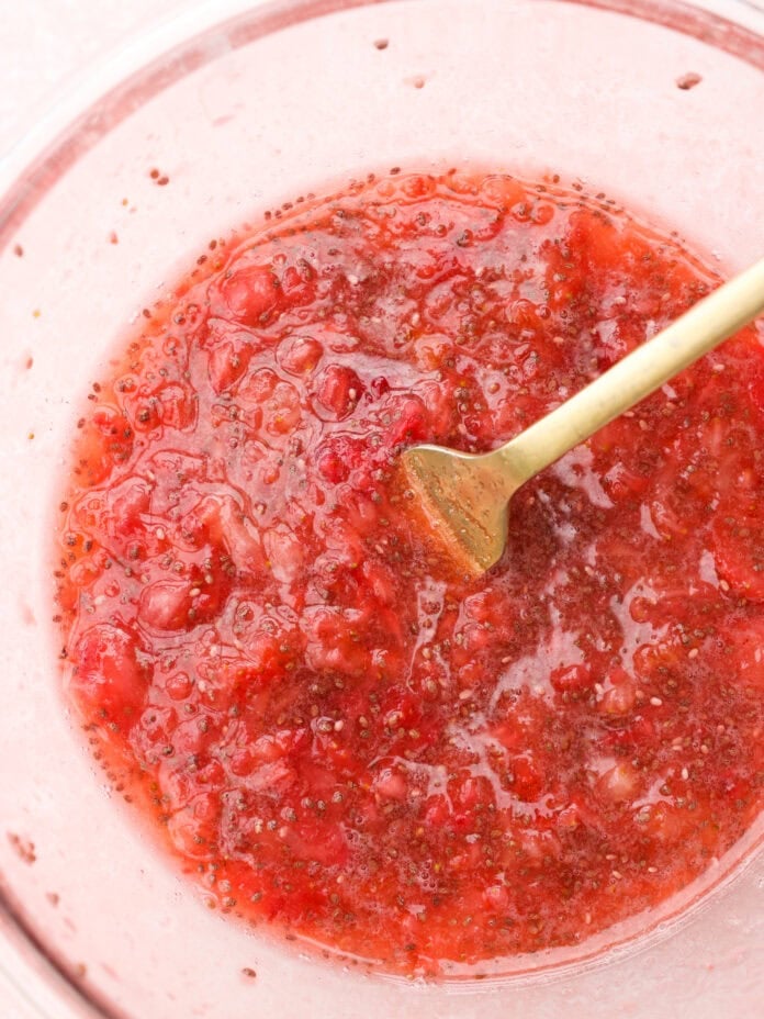 A glass bowl filled with mashed strawberries and seeds, being stirred with a gold spoon.