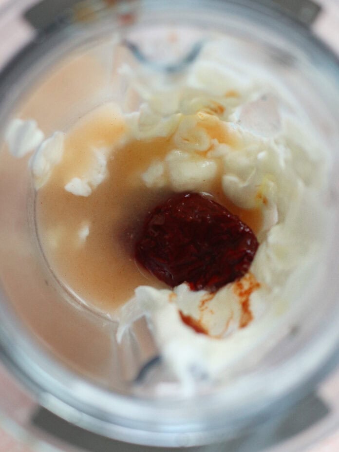 Overhead view of a blender cup containing pale liquid, pieces of chopped white onion, and a red chili pepper or paste in the center, ready to be blended.