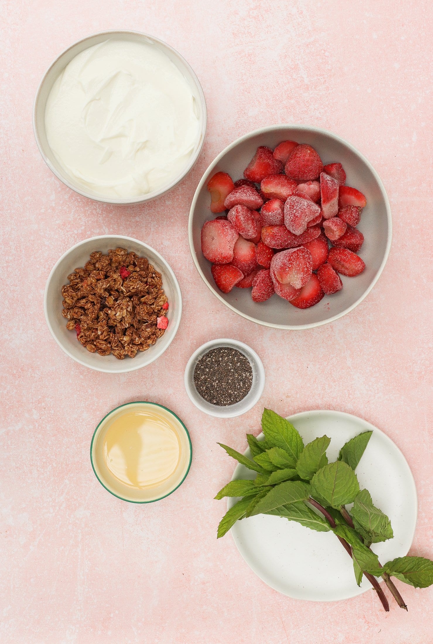 Five bowls on a pink surface contain whipped cream, frozen strawberries, granola, chia seeds, and sweetened condensed milk, next to a white plate with sprigs of fresh mint.