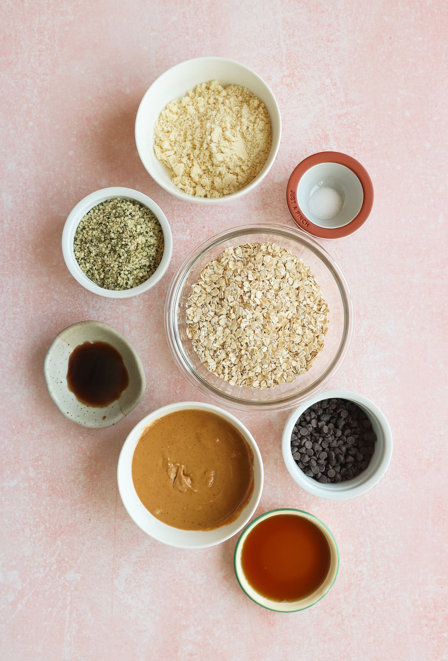 A flat lay of seven bowls on a pink surface, each containing different ingredients: oats, almond flour, hemp seeds, peanut butter, chocolate chips, vanilla extract, and maple syrup.