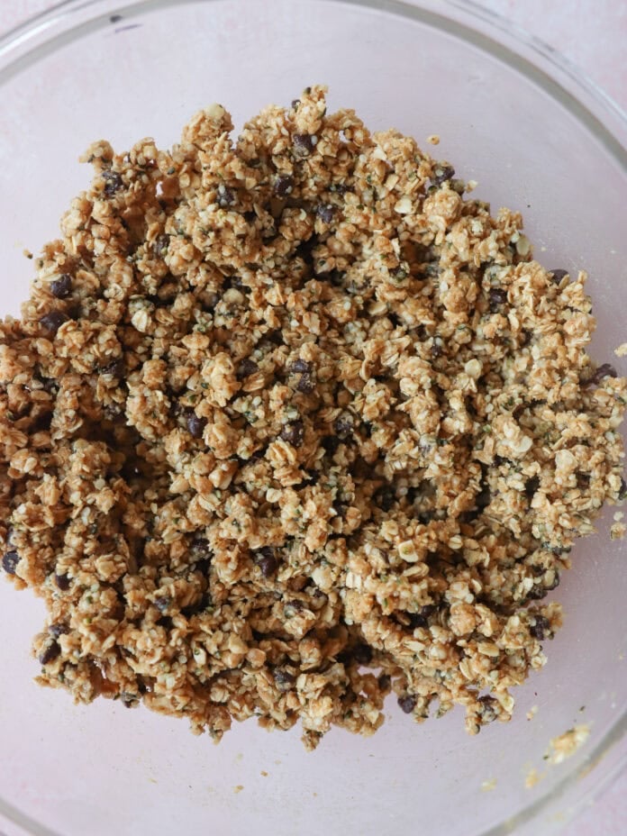 A glass bowl filled with a crumbly mixture of oats and small chocolate chips, possibly cookie or granola bar dough, sitting on a light pink surface.