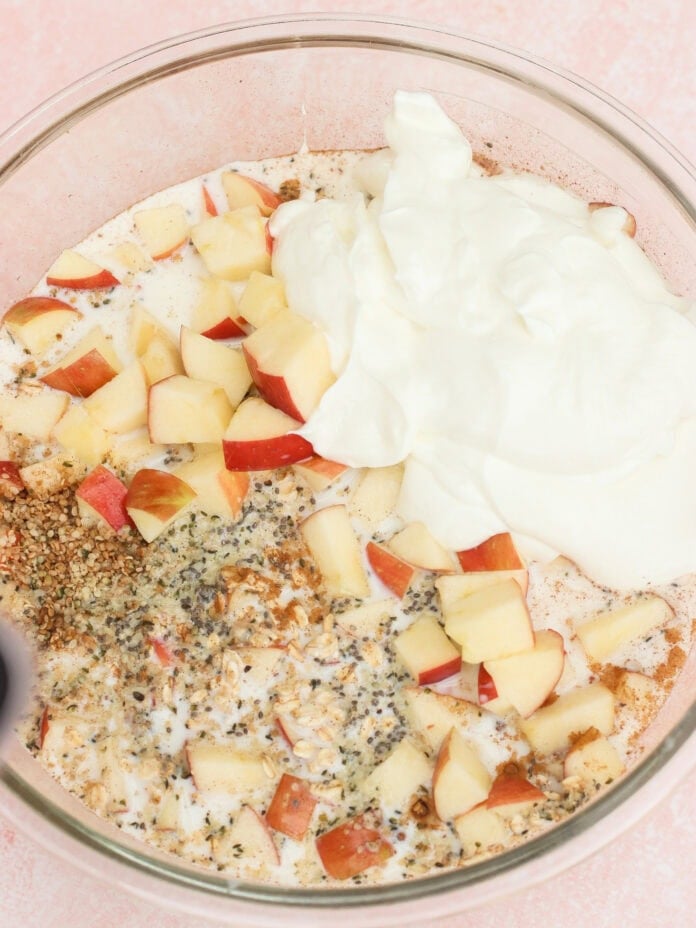 A glass bowl containing chopped apples, yogurt, seeds, spices, and milk on a pink surface, ready to be mixed for a recipe.