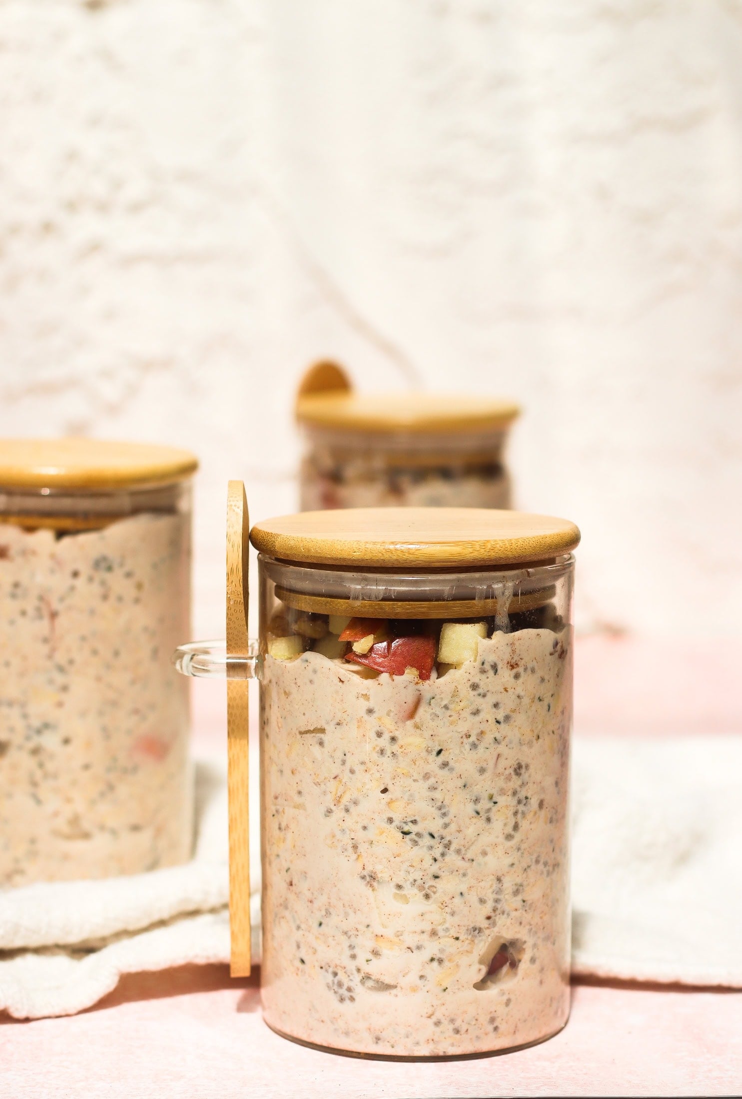 Three glass jars with wooden lids hold a creamy mixture containing visible fruit and chia seeds. The jar in the foreground is open, showing the contents more clearly. The background is softly lit and out of focus.