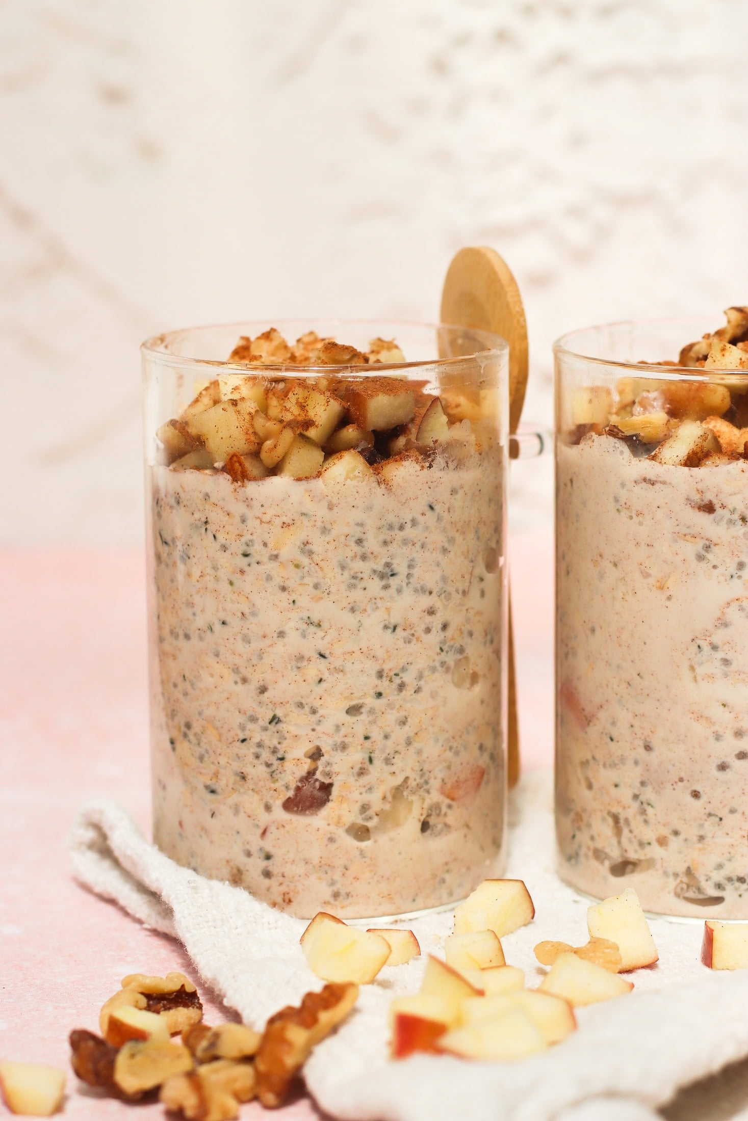 Two clear glasses filled with a creamy oatmeal mixture topped with chopped apples and walnuts. A wooden spoon rests behind one glass. Diced apples and walnuts are scattered on a white cloth beneath the glasses.