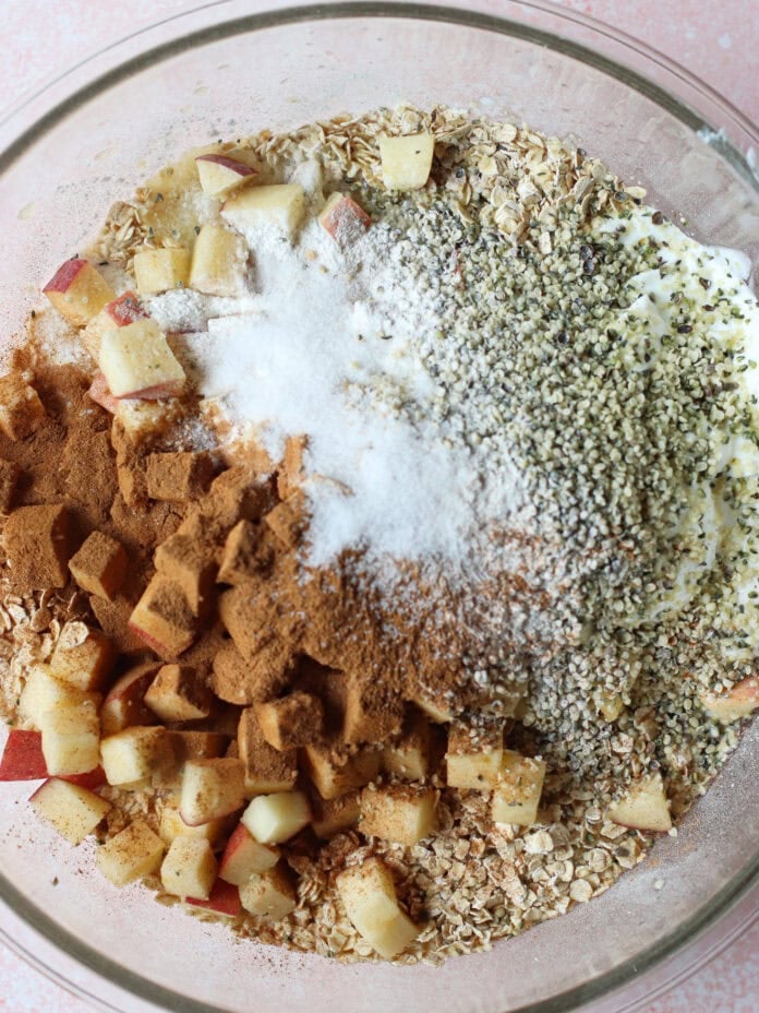 A glass bowl filled with oats, chopped apples, cinnamon, hemp seeds, yogurt, and baking powder, shown before mixing on a light pink background.