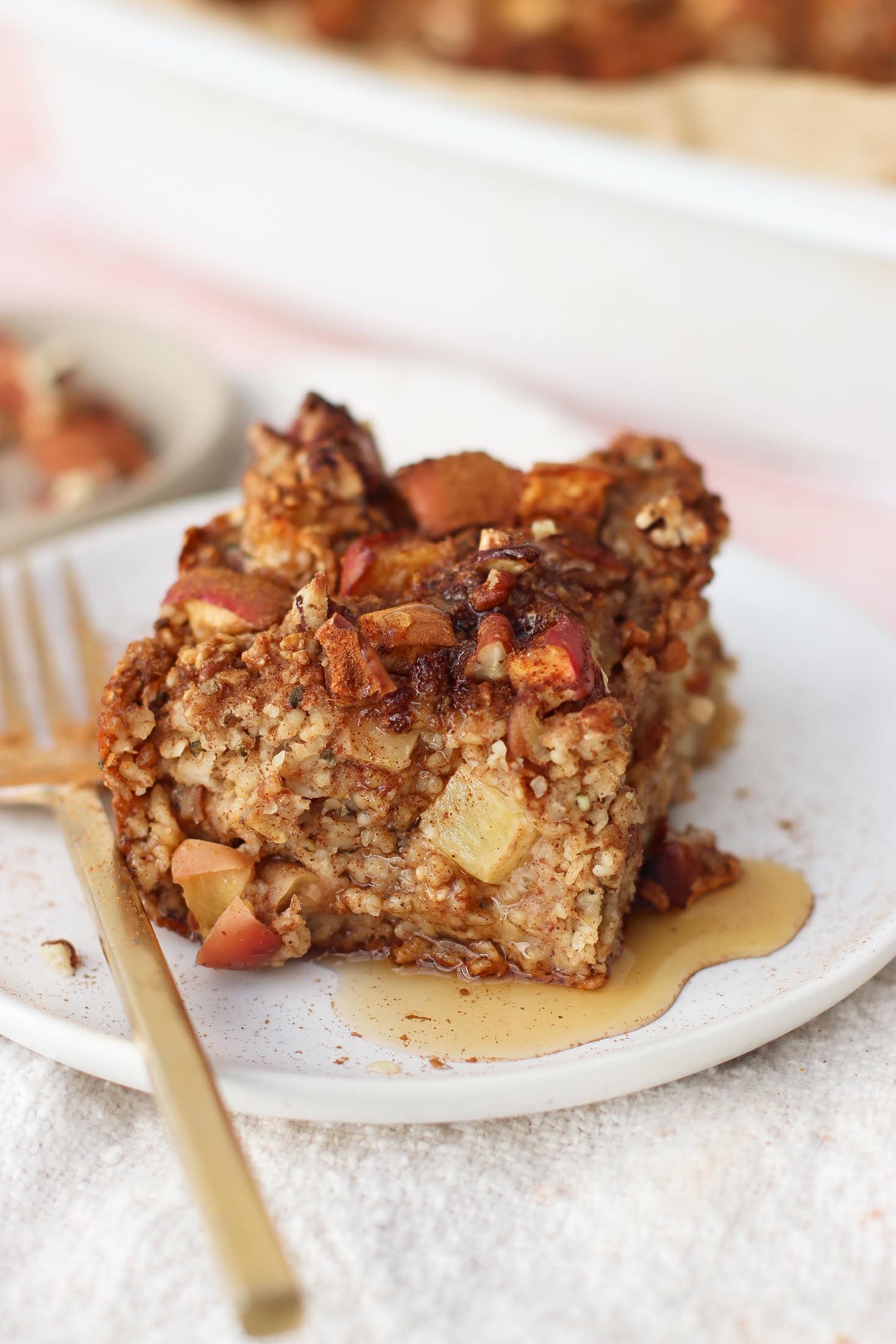 A close-up of a square slice of baked oatmeal with chopped apples, served on a white plate with a gold fork and drizzled with syrup. The background shows a baking dish and another plate with oatmeal.