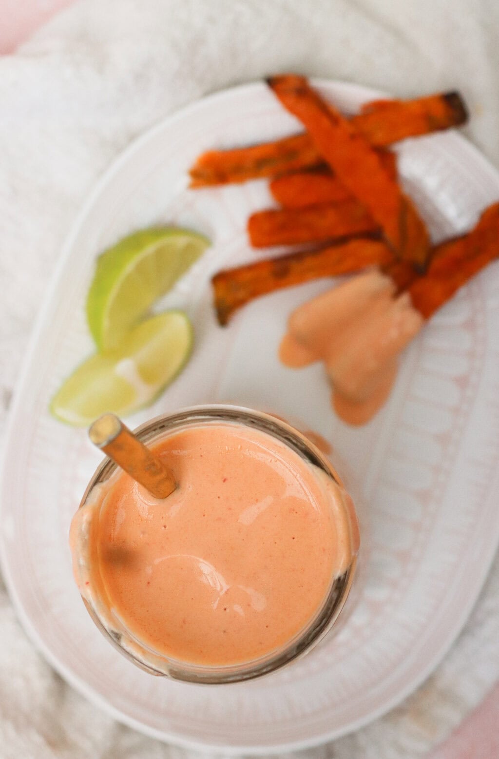 A glass of creamy pink dipping sauce with a spoon, next to sweet potato fries, two lime wedges, and a dollop of sauce on a white plate.