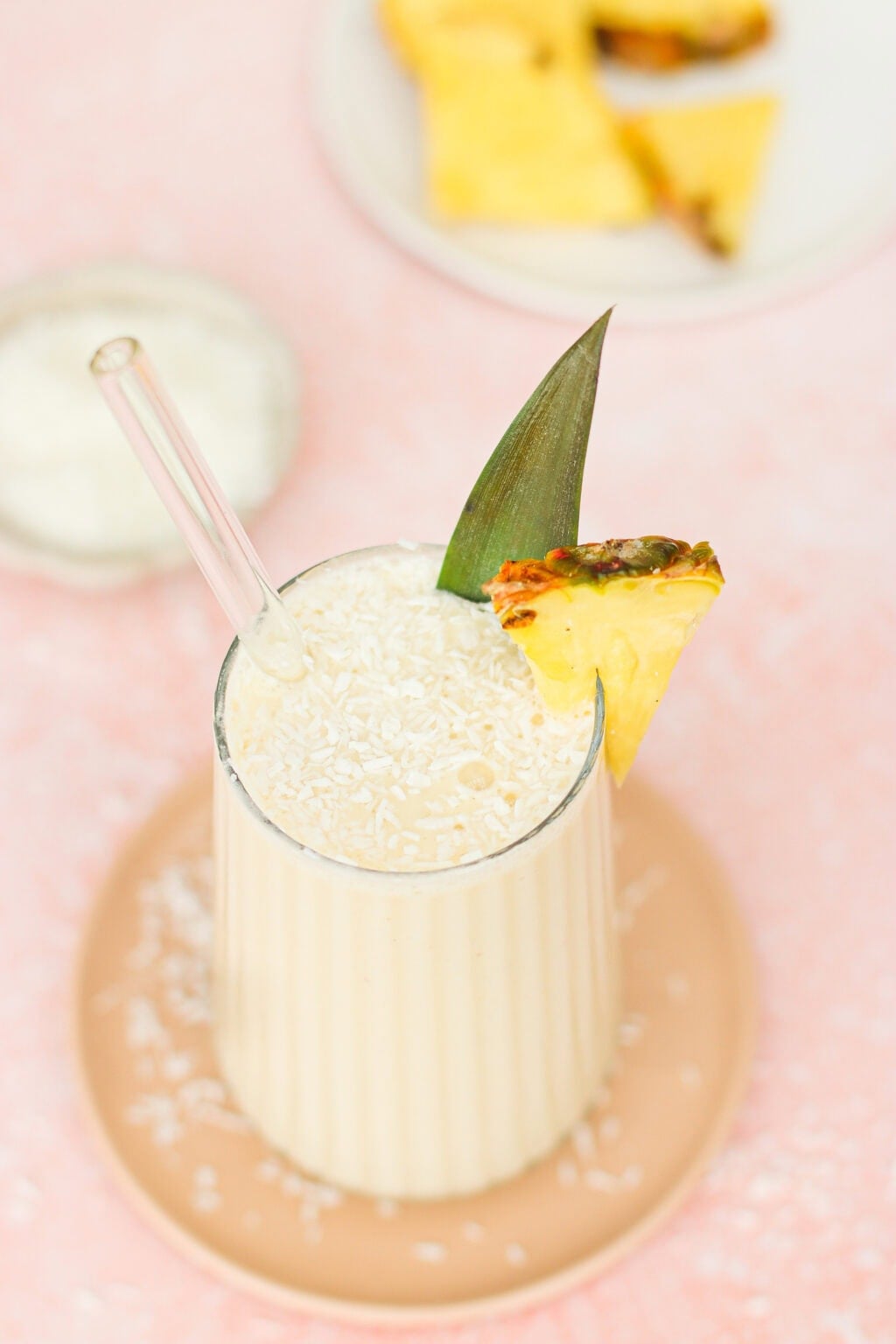 A creamy smoothie in a ribbed glass with a glass straw, topped with shredded coconut, a pineapple slice, and a pineapple leaf garnish. The drink sits on a round plate, with extra pineapple pieces in the background.