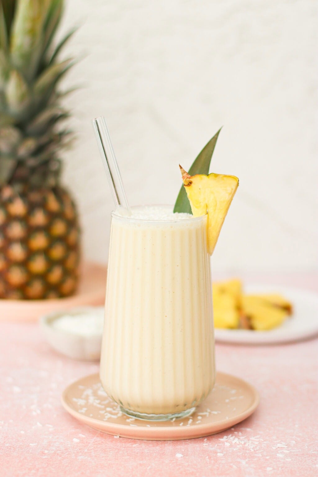 A creamy pineapple smoothie in a tall glass with a glass straw, garnished with a pineapple wedge and leaf, sits on a pink saucer. A whole pineapple and plate of pineapple slices are blurred in the background.
