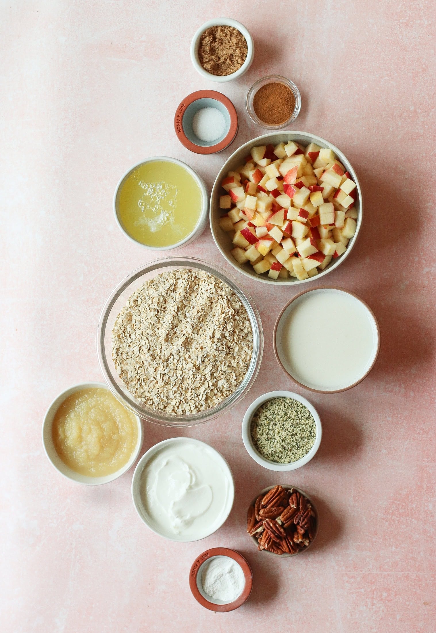 A flat lay of bowls on a pink surface containing chopped apples, oats, pecans, milk, applesauce, yogurt, melted butter, cinnamon, brown sugar, hemp seeds, and baking powder.