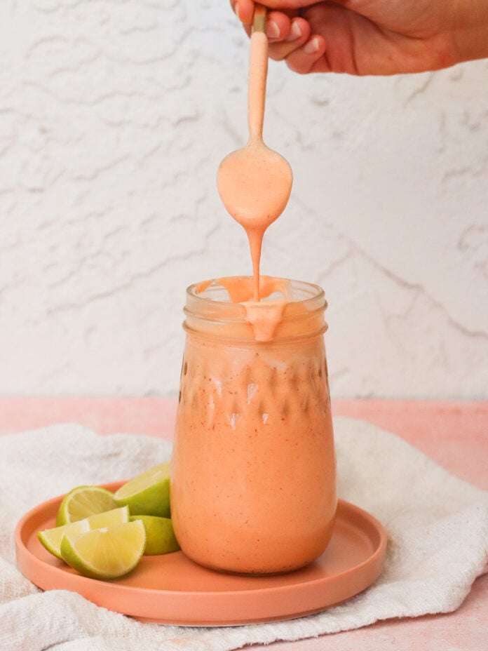 A hand holds a spoon drizzling thick, creamy, orange sauce into a mason jar on a peach-colored plate with lime wedges beside it, against a light textured background.