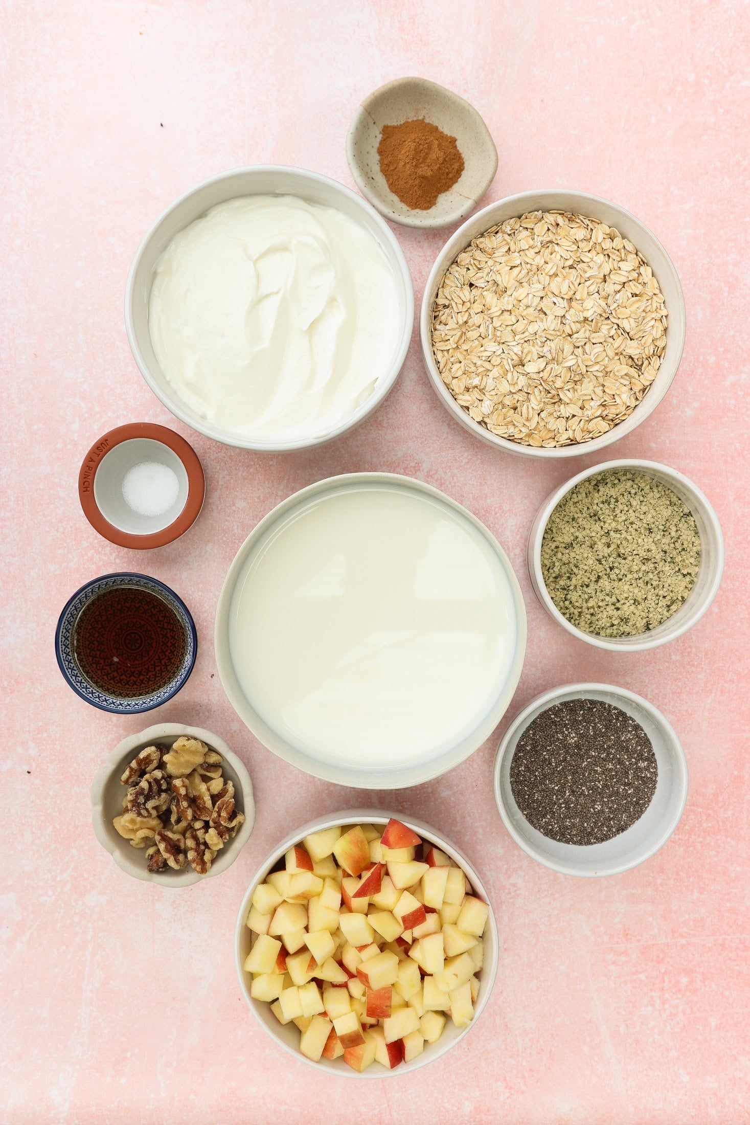 Bowls containing yogurt, rolled oats, milk, diced apples, walnuts, chia seeds, hemp seeds, maple syrup, cinnamon, and a pinch of salt, arranged neatly on a light pink background.
