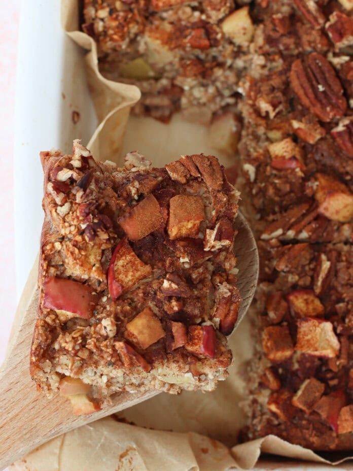 A close-up of a baked oatmeal square with apple chunks and pecans, lifted from a pan with a wooden spatula. The oatmeal is golden brown and the pan is lined with parchment paper.