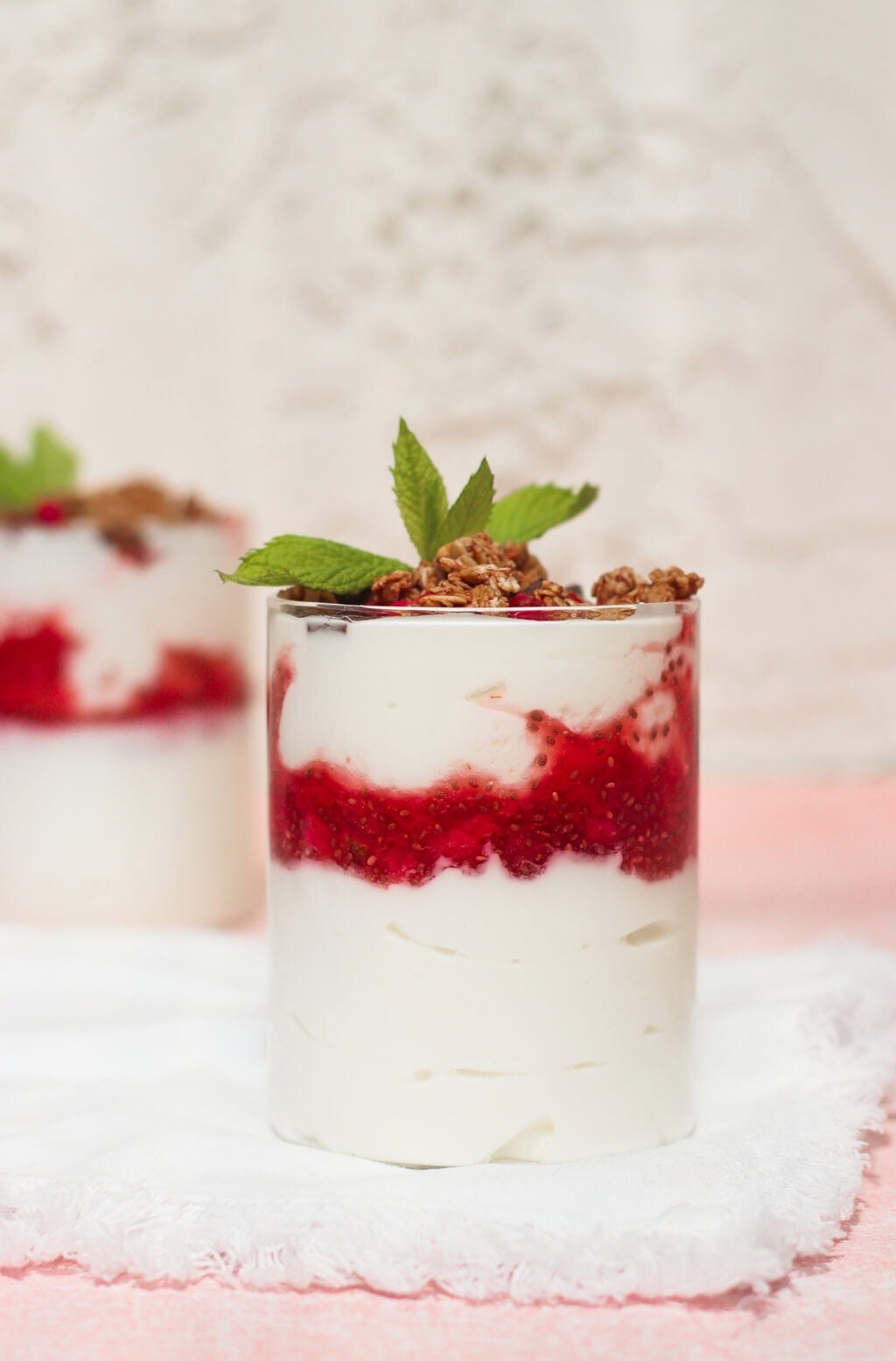 A glass filled with creamy yogurt layered with red berry sauce, topped with granola and a sprig of fresh mint, sits on a white napkin with another similar dessert blurred in the background.