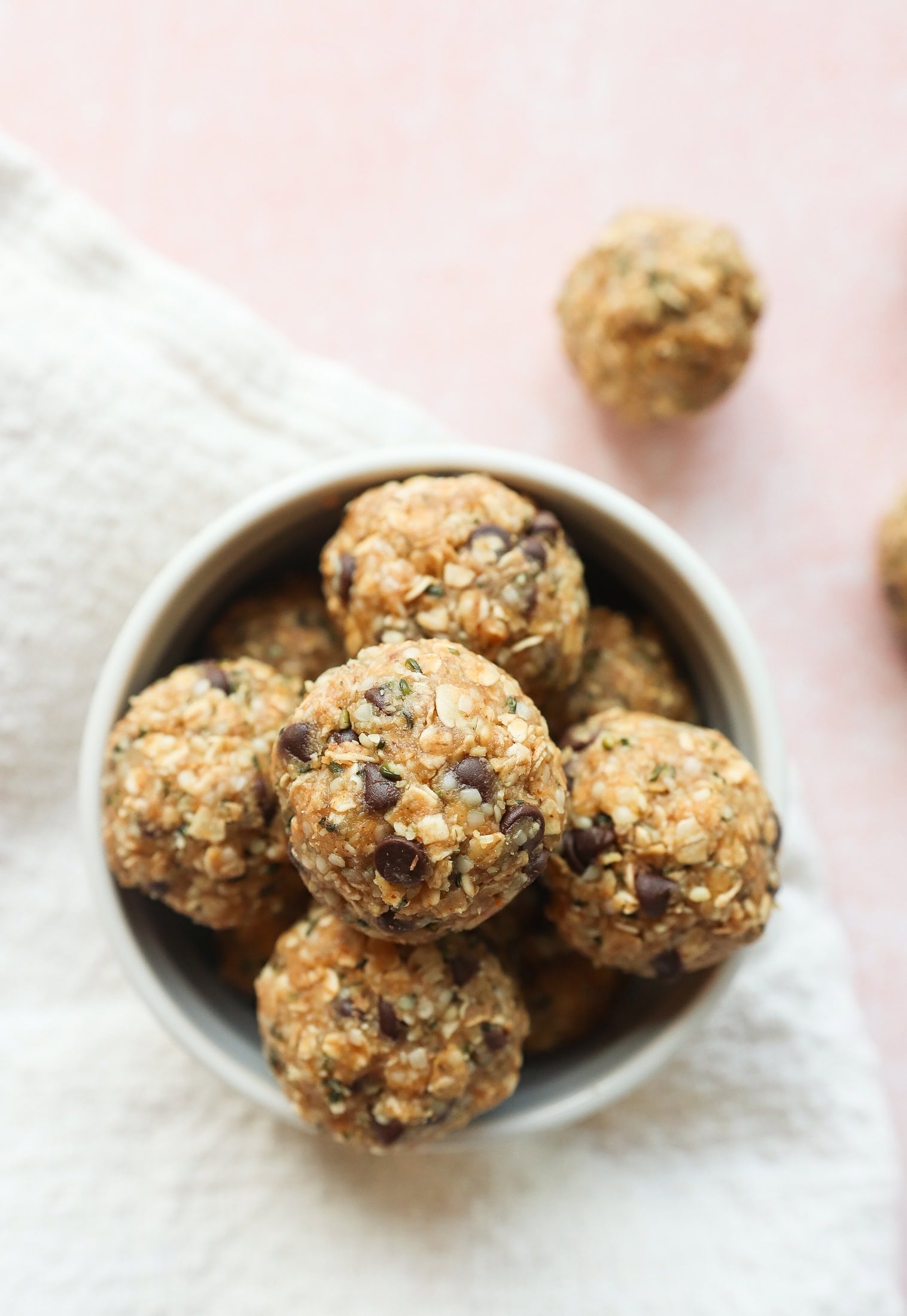 A bowl filled with round energy bites made of oats, chocolate chips, and seeds sits on a white textured cloth with a few bites scattered outside the bowl on a light pink surface.