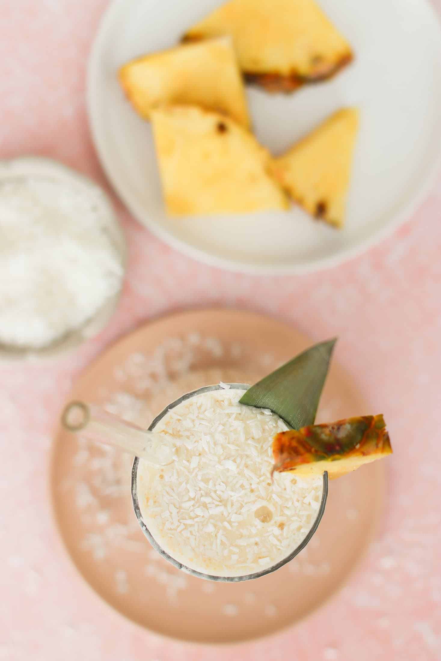 A creamy drink topped with shredded coconut and garnished with a pineapple slice and leaf, served with a glass straw. In the background, there are pineapple wedges and a small bowl of shredded coconut.