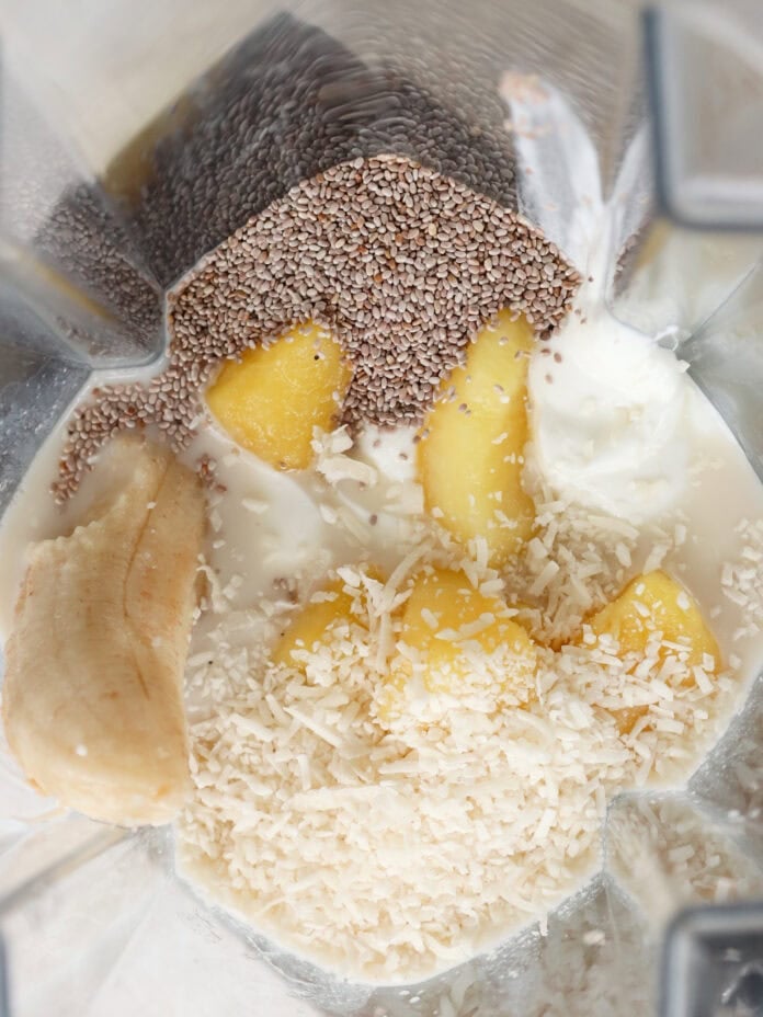 A close-up view from above of a blender containing a banana, pineapple chunks, shredded coconut, chia seeds, and yogurt, ready to be blended.