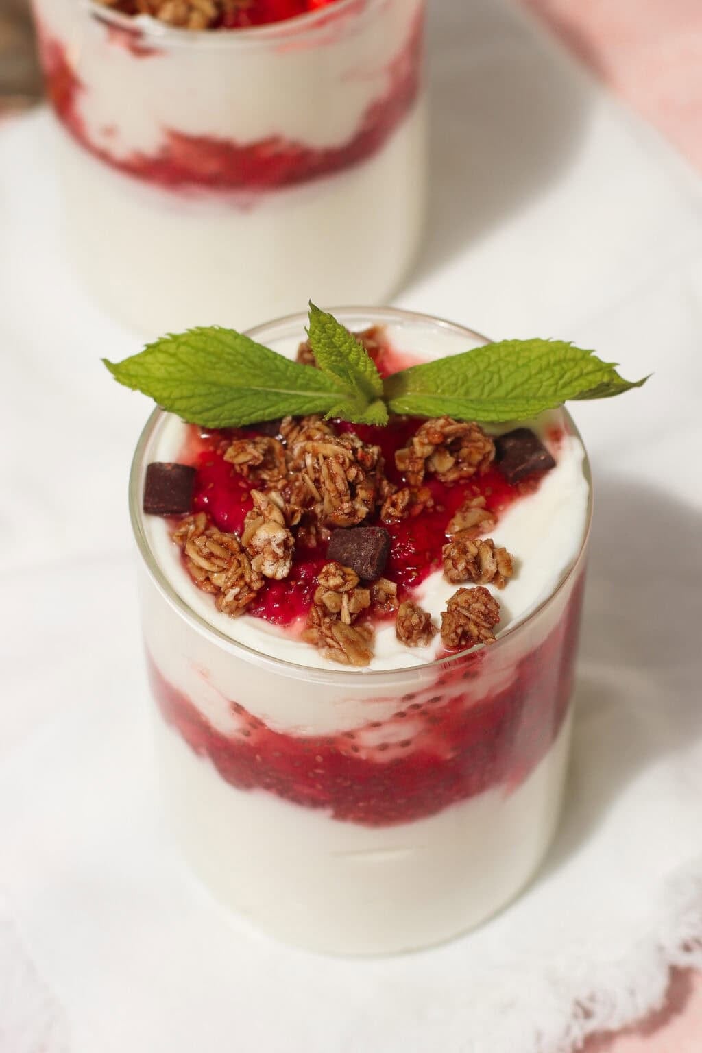 A glass of yogurt parfait layered with red berry sauce, topped with granola, chocolate chunks, and a sprig of fresh mint, placed on a white napkin.