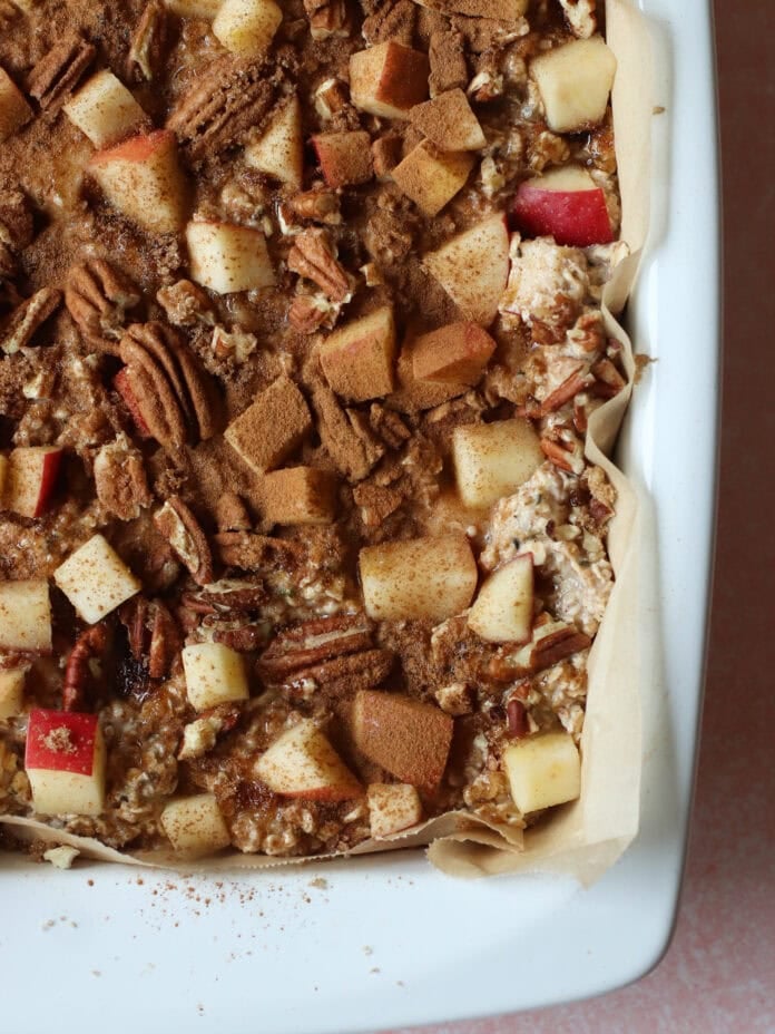 A close-up of a baking dish filled with a mixture of oats, chopped apples, pecans, and sprinkled cinnamon on top, lined with parchment paper.