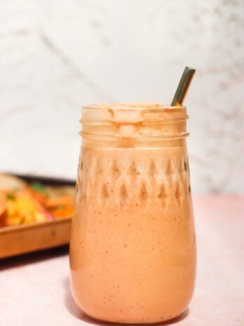 A mason jar filled with a creamy, light orange smoothie and topped with froth, with a metal straw inside. The jar sits on a light pink surface, and the background is softly blurred.