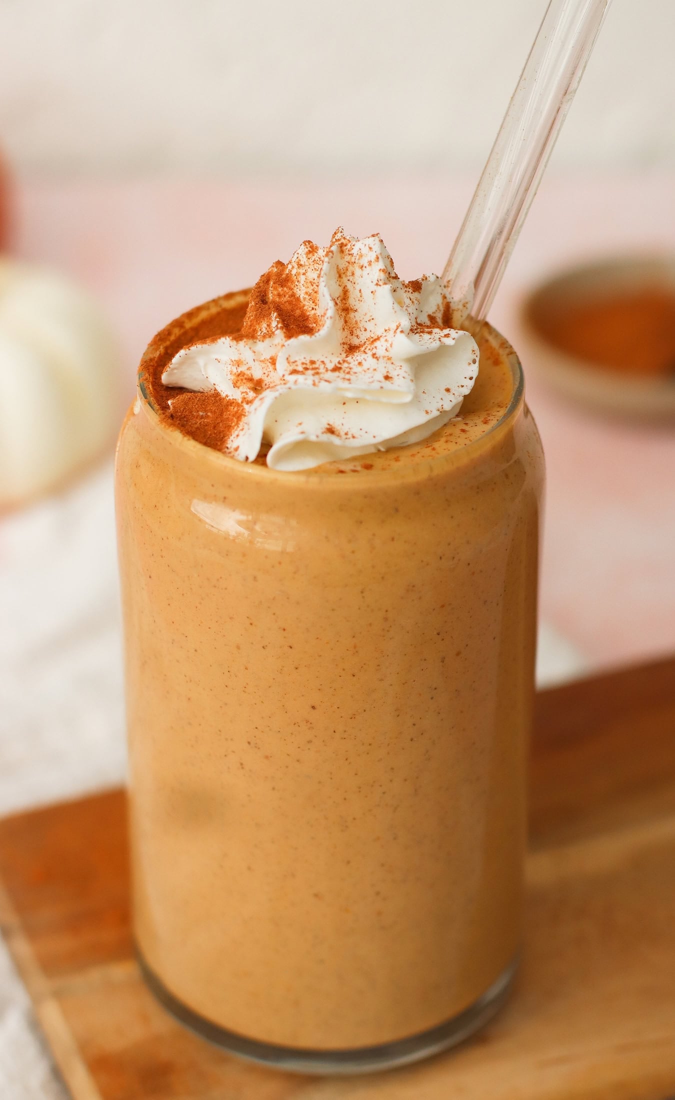 A glass filled with a creamy, brown smoothie topped with whipped cream and a sprinkle of cinnamon, with a clear straw inserted, sitting on a wooden surface.