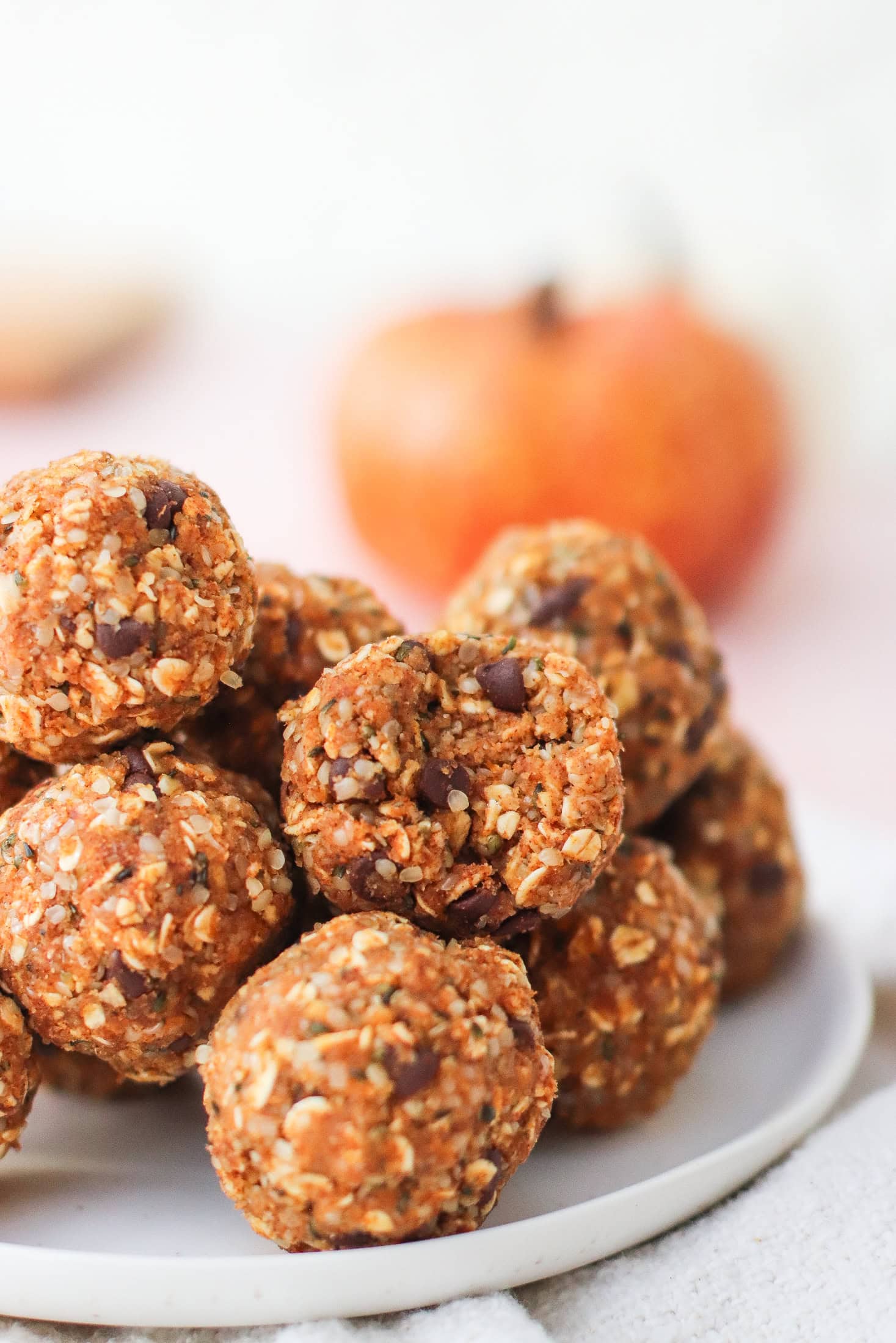 A stack of oat and chocolate chip energy bites sits on a white plate, with a blurred orange pumpkin in the background.